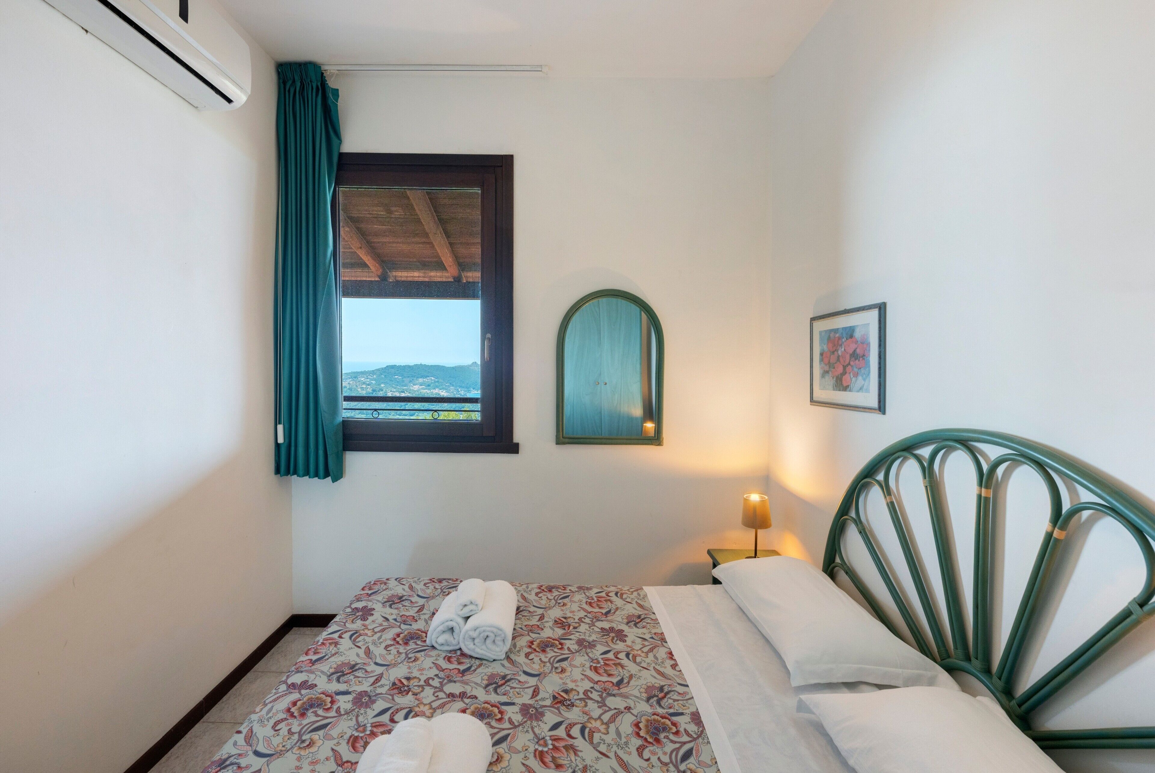 Un dormitorio con cama, ventana con vistas, lámpara de noche y un cuadro en la pared.
