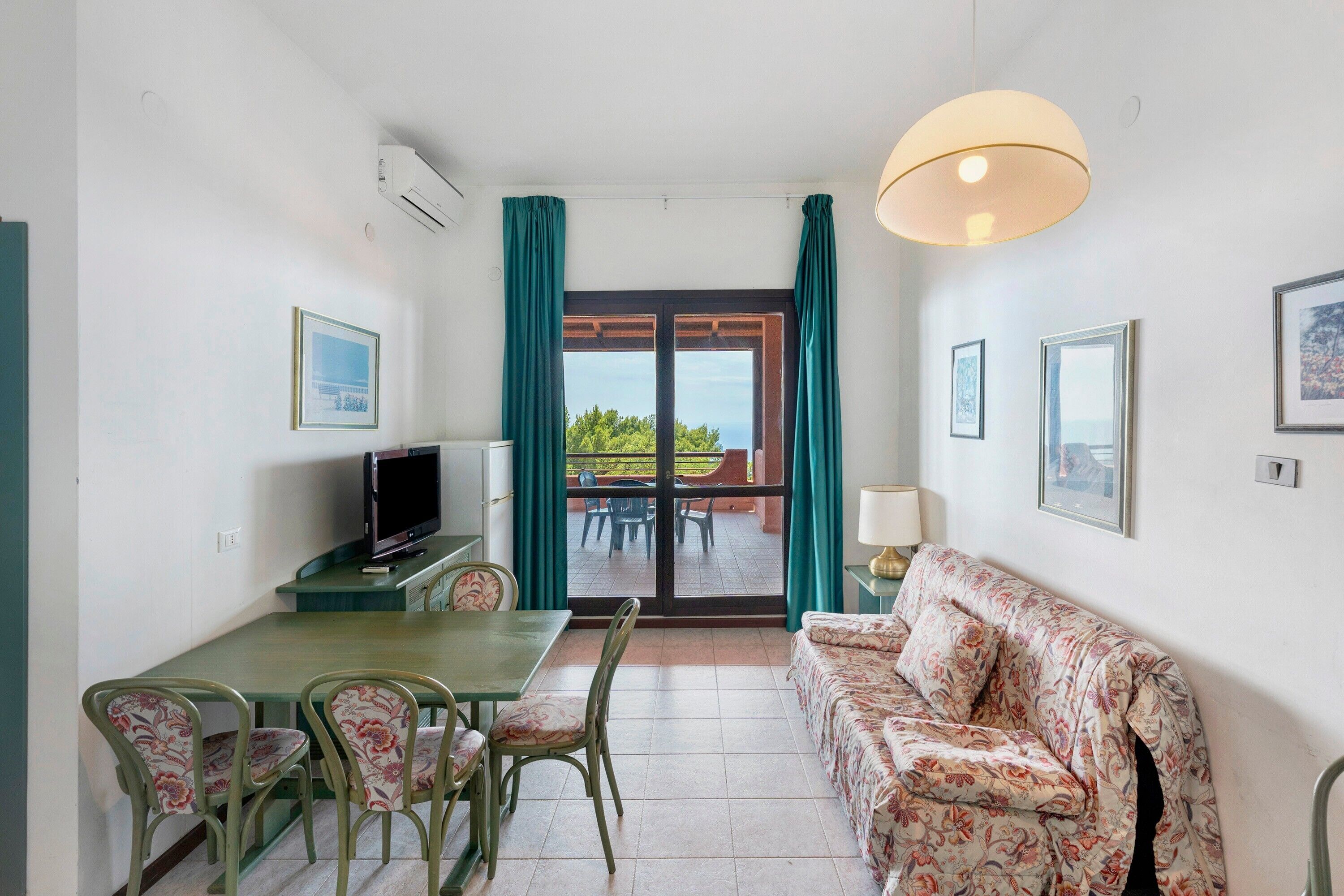Une pièce comprenant une table à manger, des chaises, un canapé, un téléviseur et une vue sur l’extérieur, visible à travers une porte vitrée.