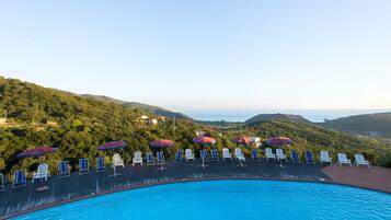 Un espace aménagé au bord de la piscine, avec des chaises longues et des parasols, entouré d’une végétation luxuriante et de collines.