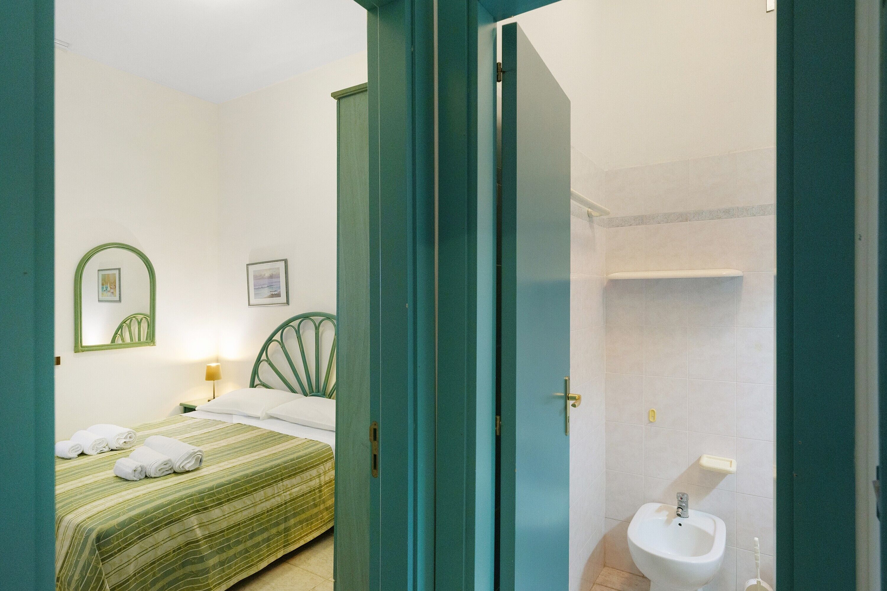 Una habitación de hotel con cama, mesita de noche con lámpara, y baño con lavabo e inodoro.