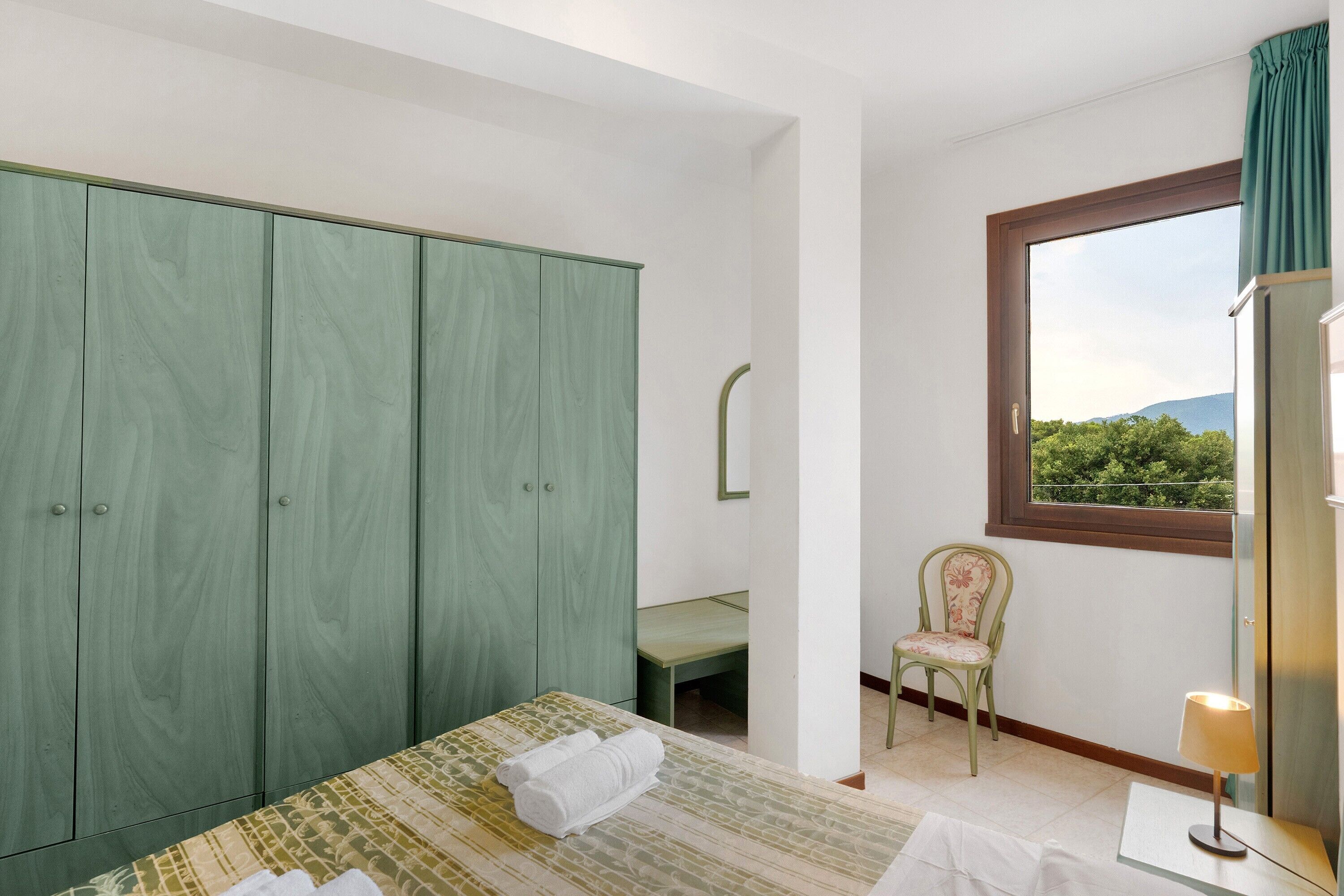 Ein Schlafzimmer mit einem Bett, einem grünen Kleiderschrank, einem Sessel, einem Fenster mit Ausblick und einem Nachttisch mit Lampe.
