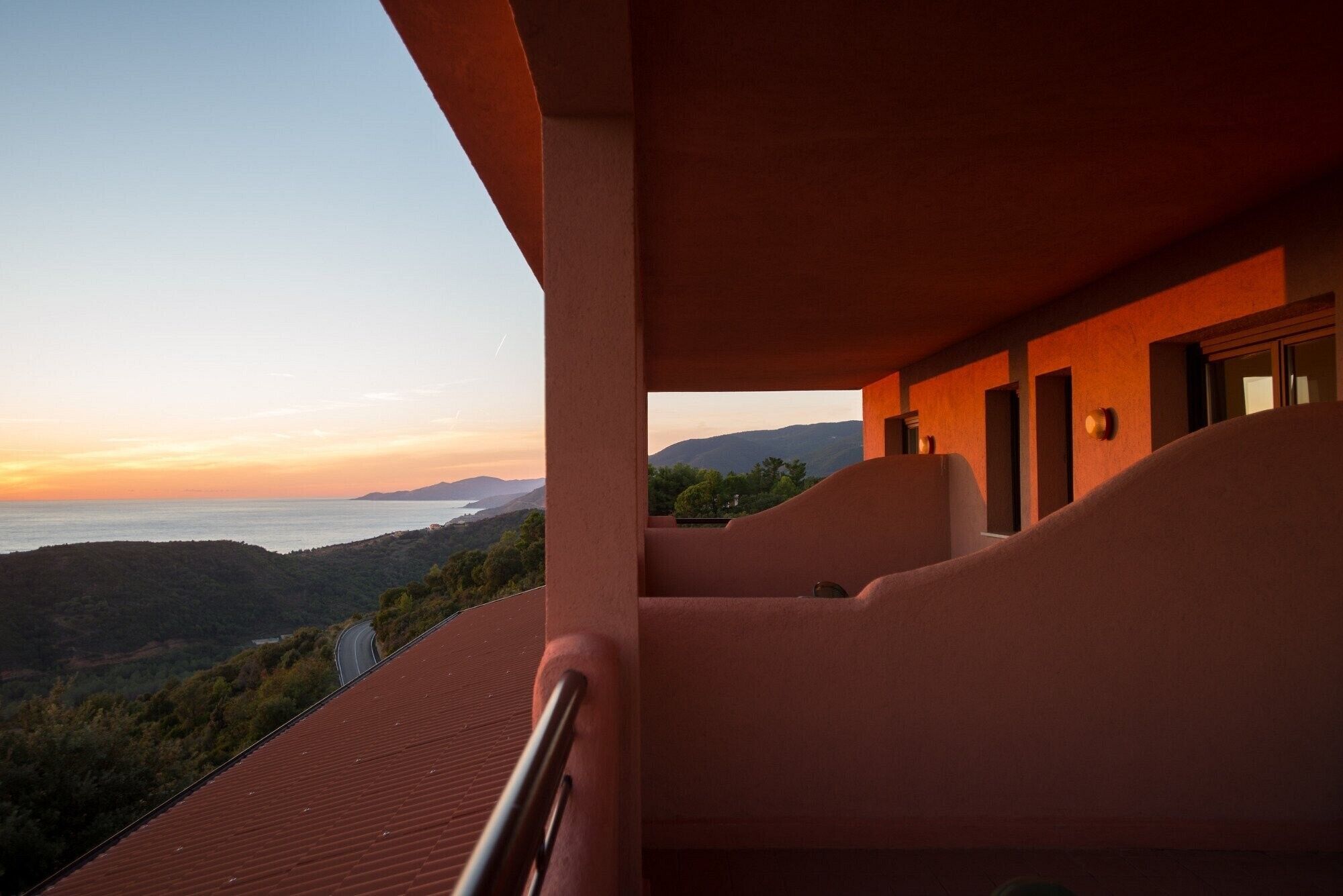 A balcony with a view of the sea and hills, a red roof, and a building with a curved facade.