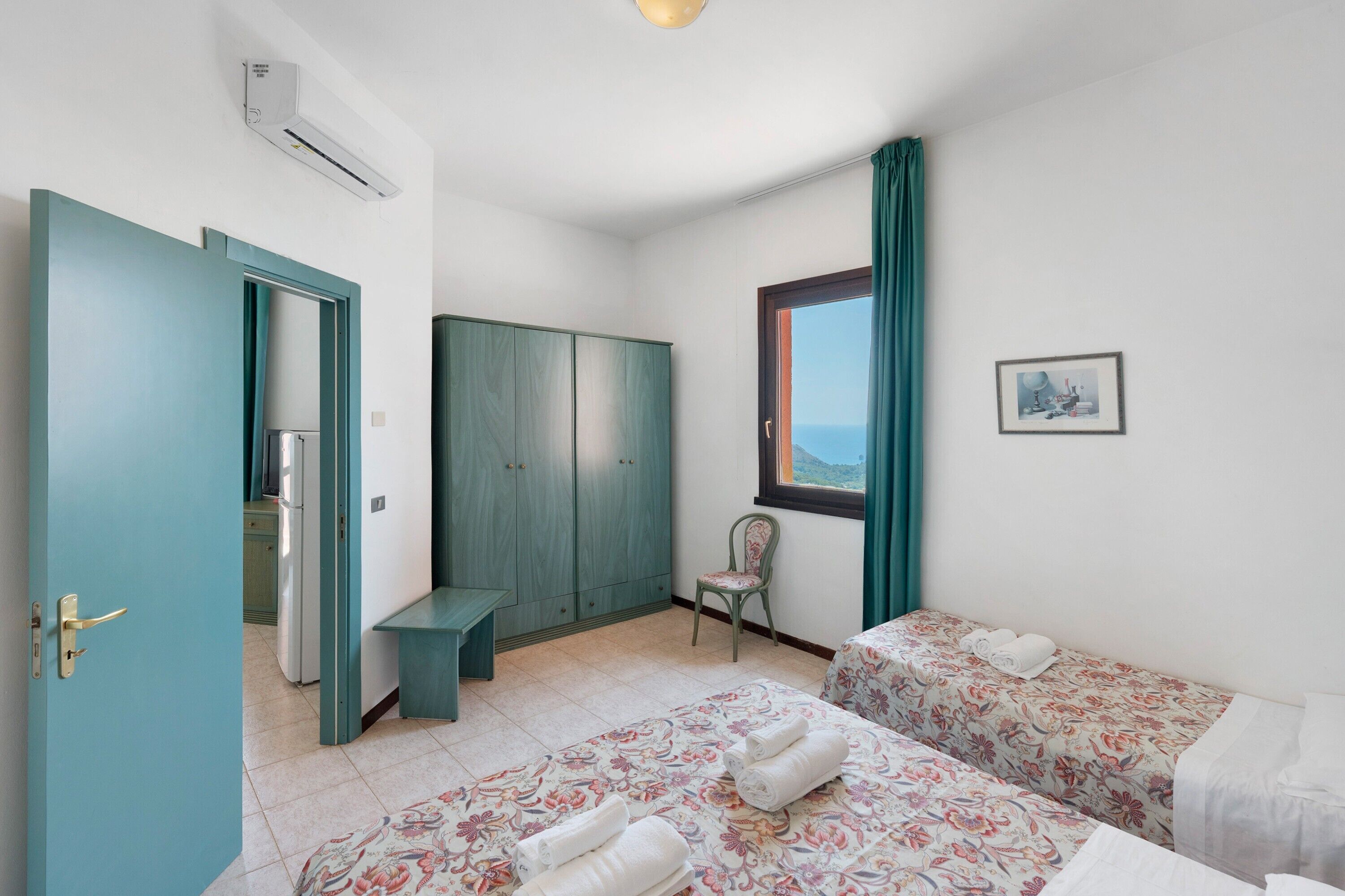 A bedroom with two beds, a wardrobe, a chair, a window with curtains, and an air conditioning unit.