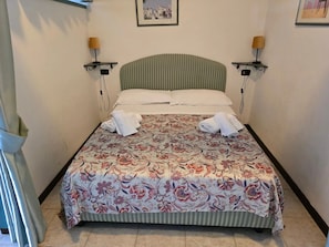 Un lit bien fait, avec une literie à motifs floraux, deux lampes fixées au mur et un tableau encadré au-dessus du lit.