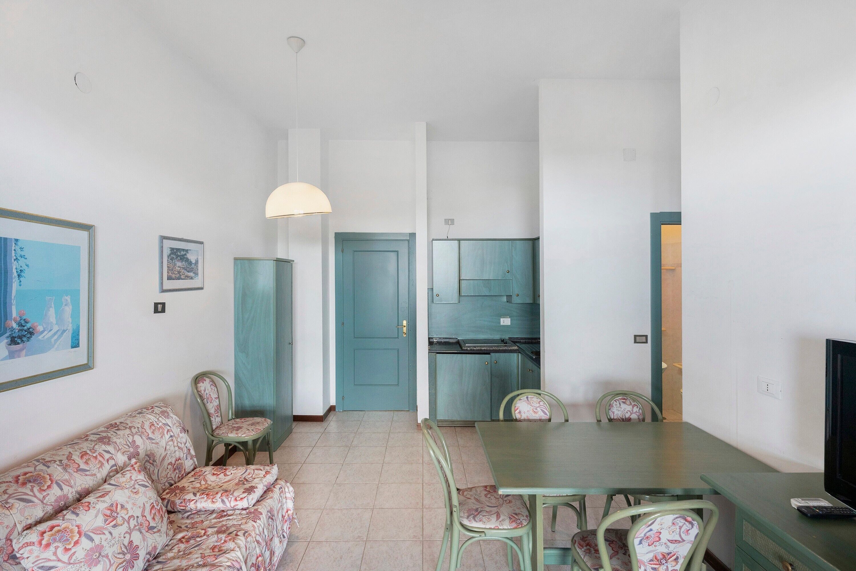 A living room with a sofa, chairs, a dining table, a TV, and a kitchen area with cabinets and a door.