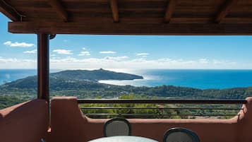Ein Balkon mit Tisch und Stühlen, mit Blick auf eine Küstenlandschaft mit Hügel und Meer.
