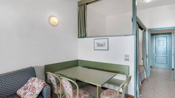 Una habitación con un banco verde, una mesa y cuatro sillas con tapicería floral. Hay un cuadro en la pared y una puerta al fondo.