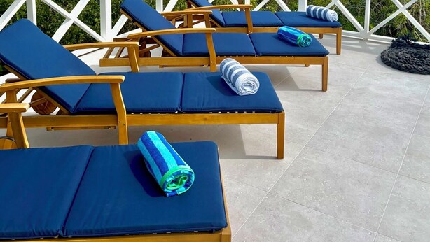 A deck with wooden lounge chairs and blue cushions, overlooking a turquoise sea and a clear sky. - Luxury Simplicity On The Beach, Little Exuma.  Brand new, 2 bedrooms. (little exuma, Bahamas)