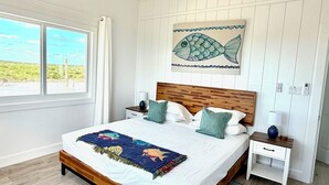 A bedroom with a wooden bed, a ceiling fan, a nightstand, a lamp, and a fish-themed artwork on the wall. - Luxury Simplicity On The Beach, Little Exuma.  Brand new, 2 bedrooms. (little exuma, Bahamas)