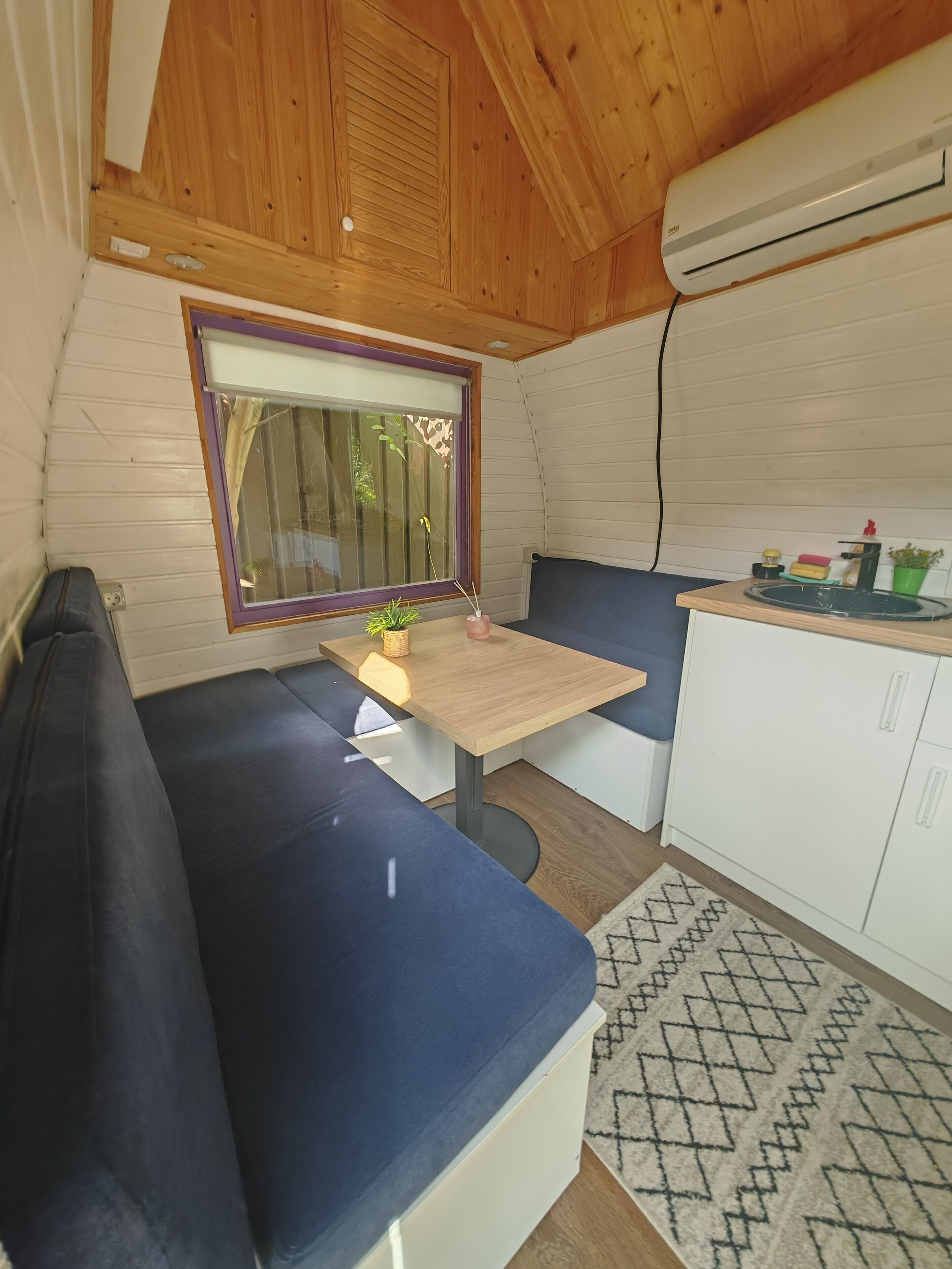 A compact kitchen with a wooden ceiling, a window with a purple frame, a small table, and a blue bench.