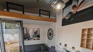 Un espace de vie moderne avec un canapé gris, une table à manger avec des chaises, une horloge murale et une grande fenêtre avec des stores.