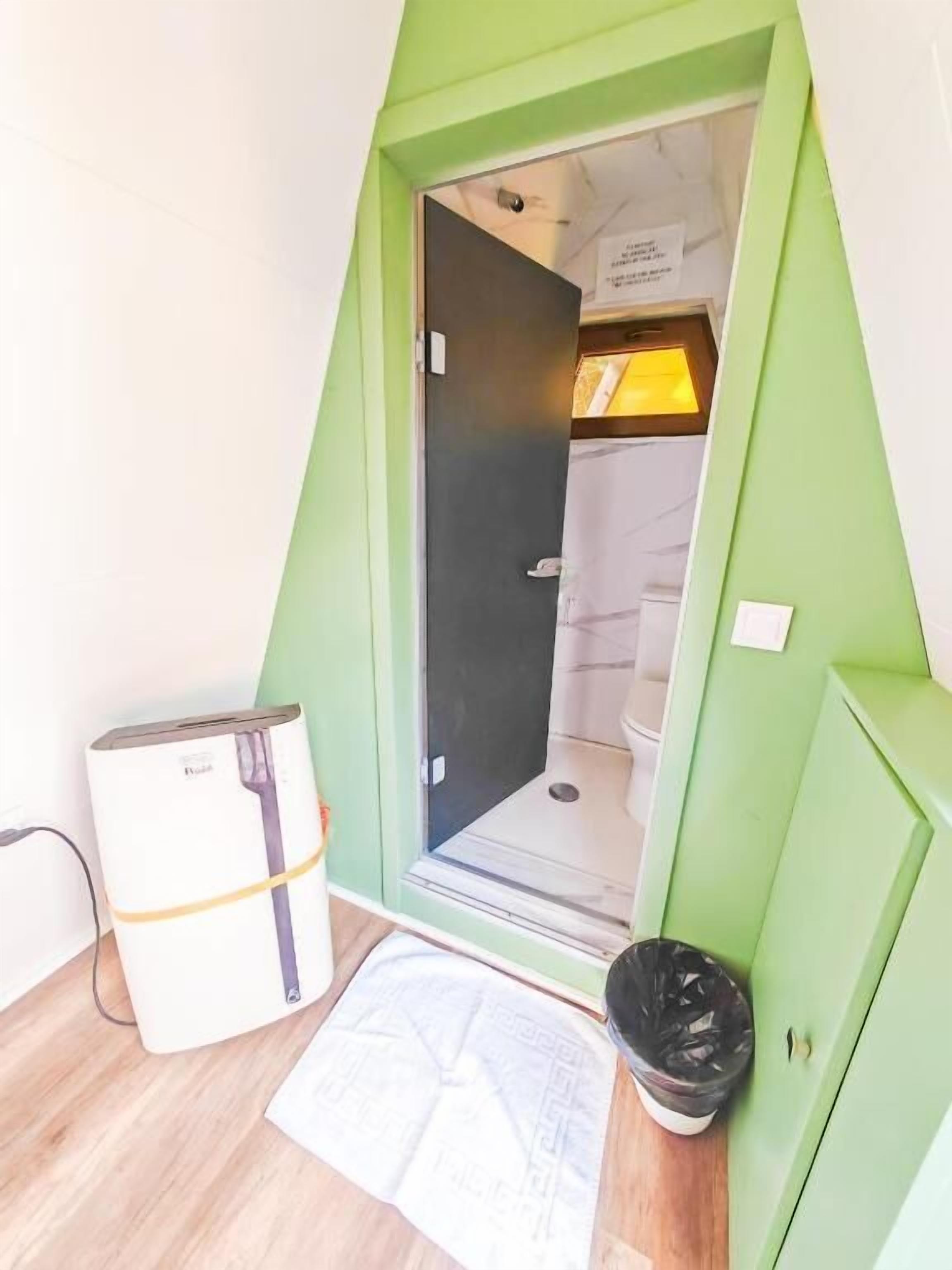 Un baño pequeño con forma triangular que incluye una ducha, un inodoro y un lavamanos. Hay un cubo de basura y un calentador pequeño en el suelo.