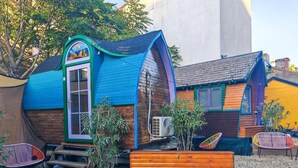 Une maison en bois aux couleurs vives, avec un toit bleu, une terrasse en bois et un petit jardin agrémenté de plantes et de meubles d’extérieur.