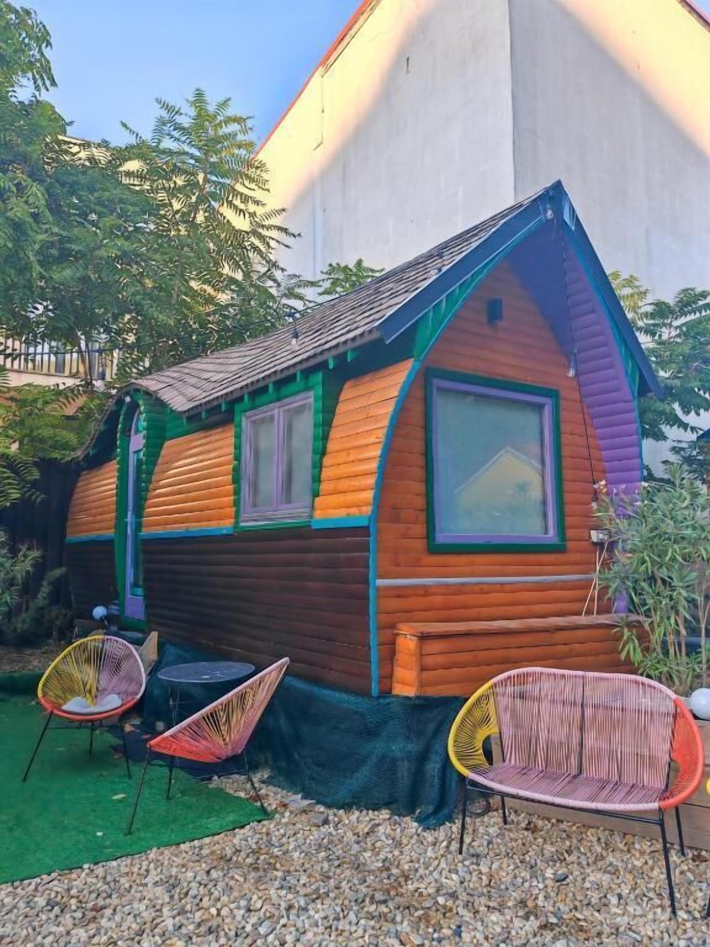 Eine farbenfrohe Holzhütte mit Satteldach, zwei Fenstern und einer kleinen Veranda mit zwei Korbstühlen.