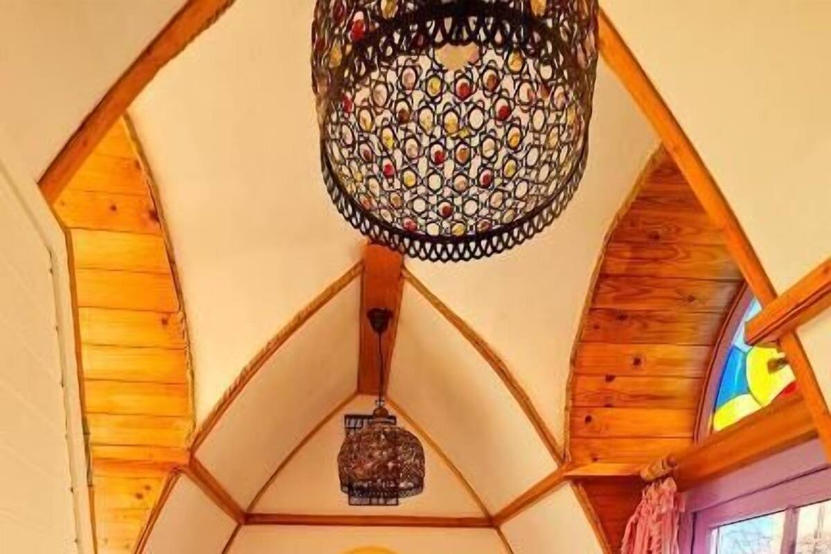 Une chambre mansardée chaleureuse, avec un plafond en pente, un lit recouvert d’une courtepointe à motifs, une petite table de chevet, un luminaire suspendu et une fenêtre avec des rideaux.