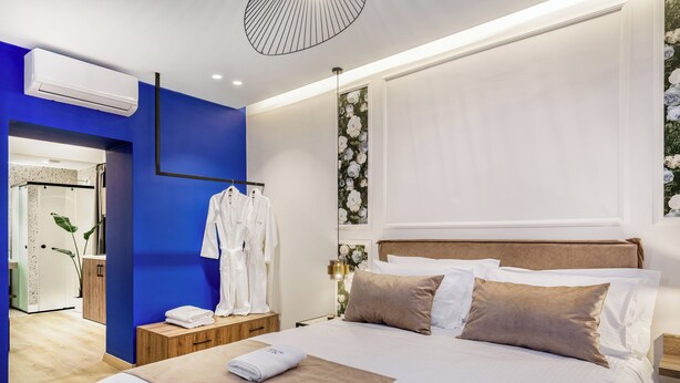 Camera d'albergo moderna con un letto grande, un comodino in legno, una parete di colore blu e un bagno visibile tramite una porta aperta.