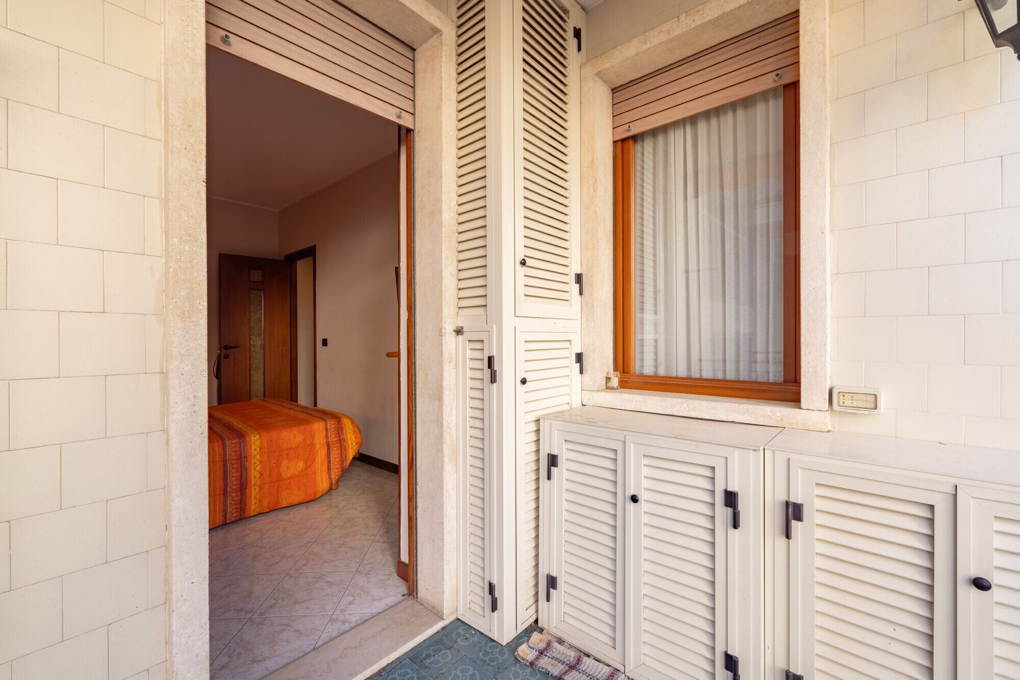 A narrow hallway with a bed at the end, a window with blinds, and a door leading to another room.