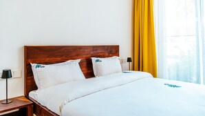 Een hotelkamer met een houten hoofdeinde, witte beddengoed, een nachtkastje met een lamp en een raam met gele gordijnen.