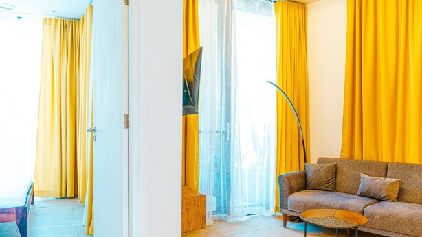 Een kamer met een bank, een klein rond tafeltje en gele gordijnen.