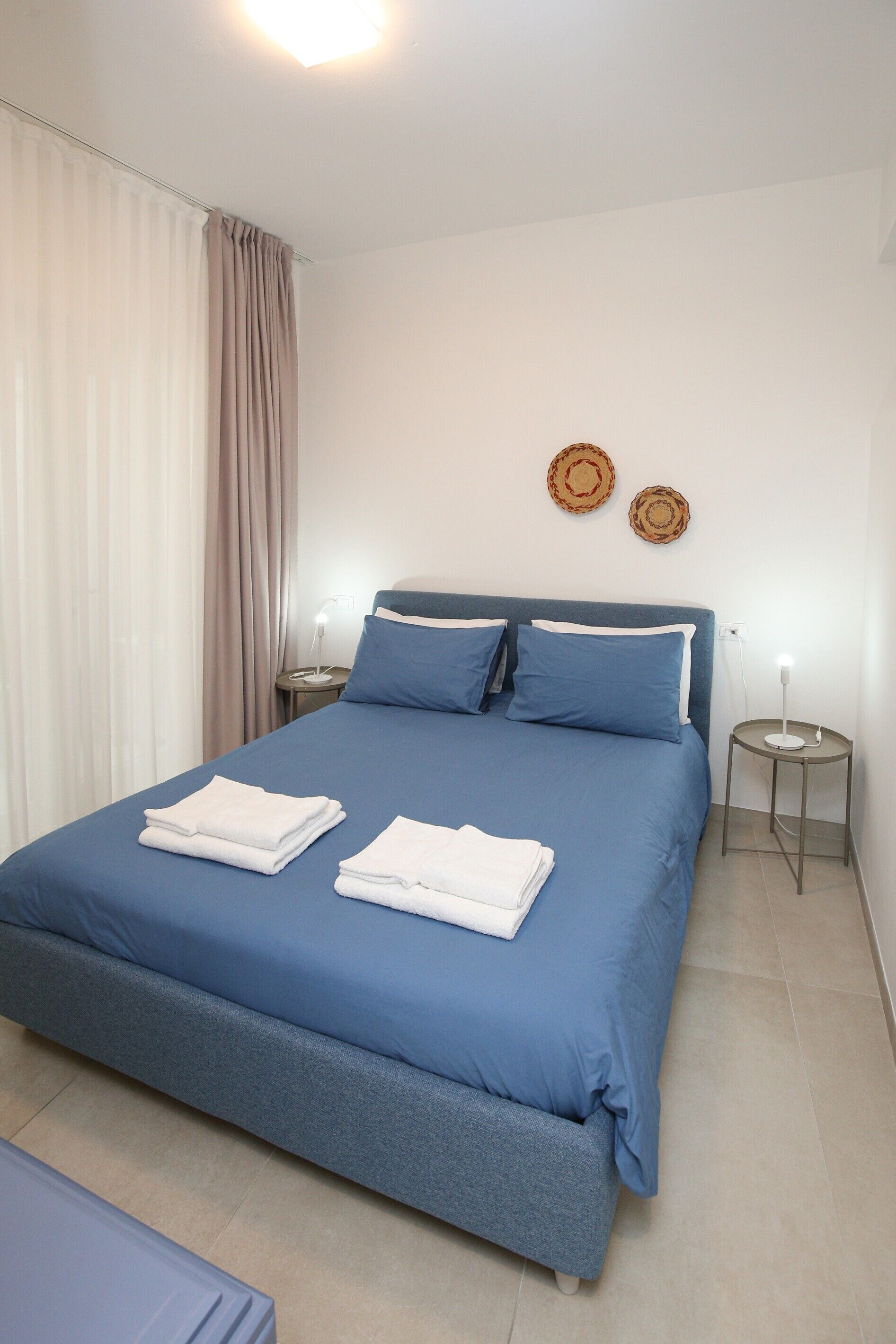 A neatly made bed with blue bedding, two bedside tables with lamps, and a curtain on the window.