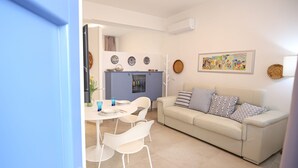 A living room with a sofa, a dining table with chairs, a kitchen area, and a wall with decorative plates and a painting.