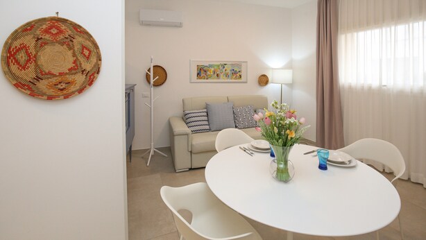 A modern living room with a round dining table, white chairs, a sofa with patterned cushions, a standing floor lamp, and a wall-mounted woven artwork.