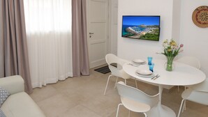 A modern living room with a white sofa, a dining table with chairs, a flat-screen TV mounted on the wall, and a window with curtains.
