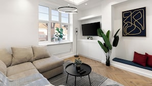 A modern living room with a sectional sofa, a round coffee table, and a large window with a view of buildings. - Apartment - Sleeps 8 - Golders Green 10 Mins (London)