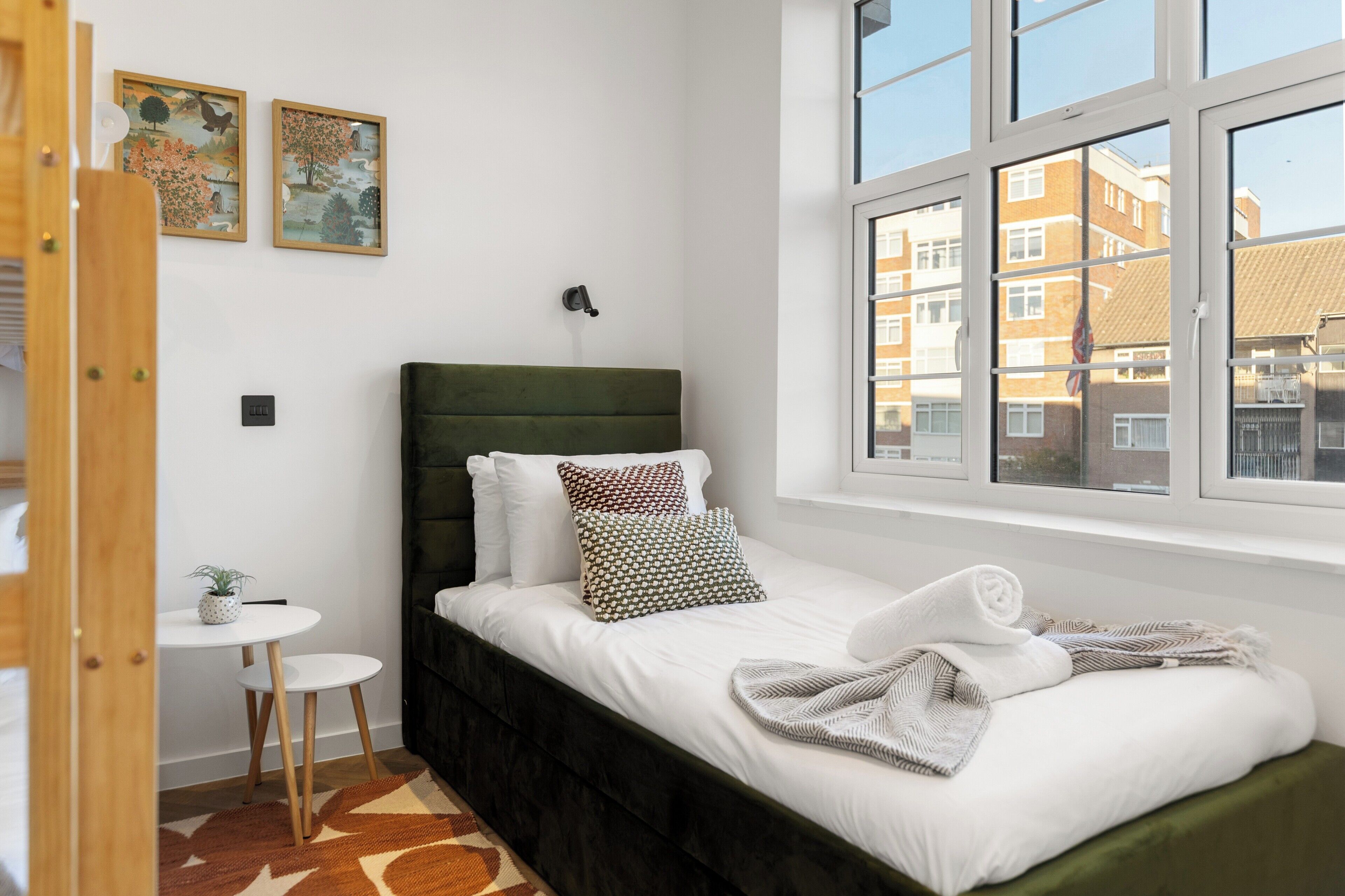 A neatly arranged bedroom with a bed, a small table with a plant, and two framed pictures on the wall.