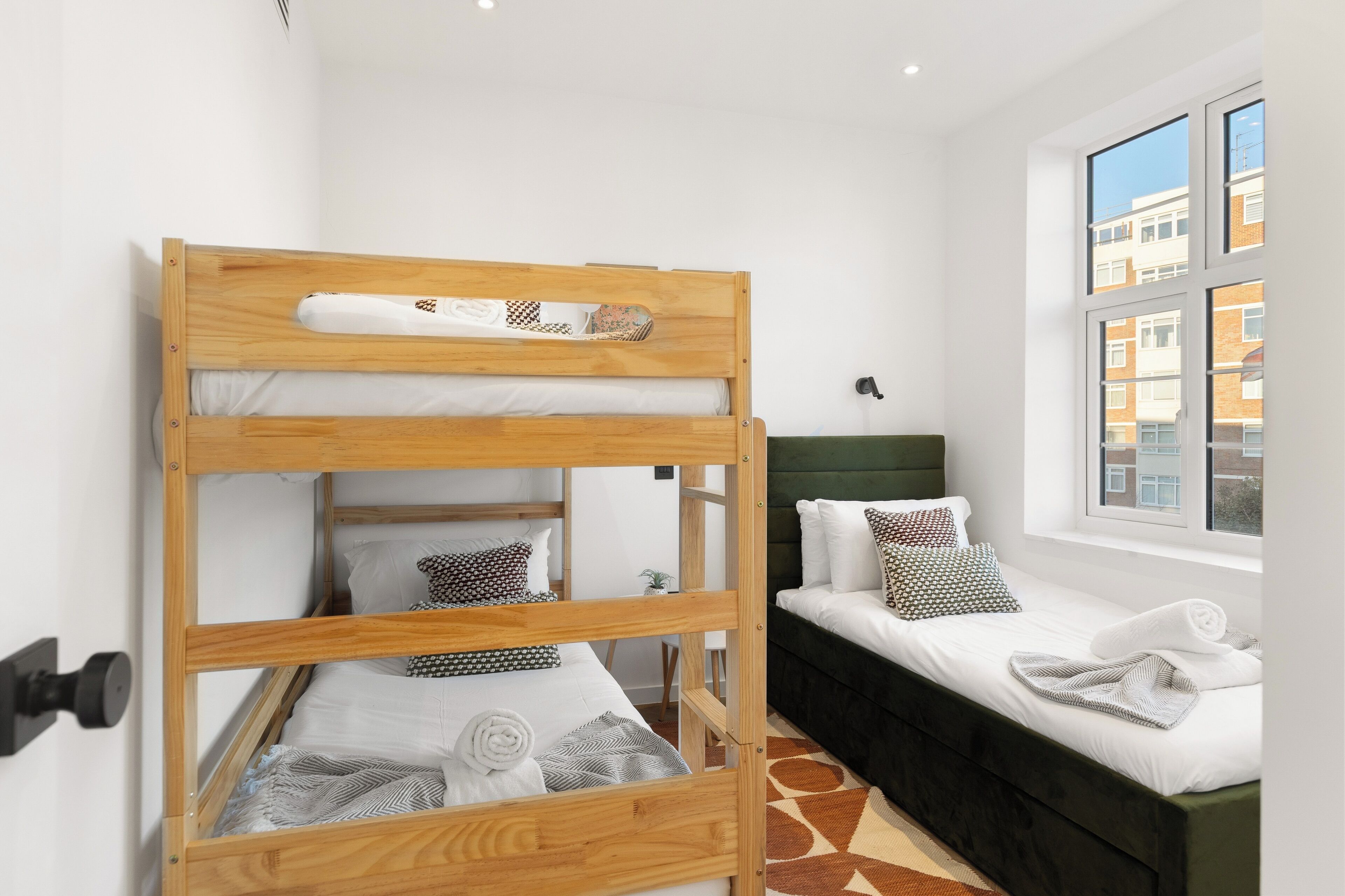 A bunk bed with a wooden frame, a single bed with a green headboard, and a window with a view of buildings.