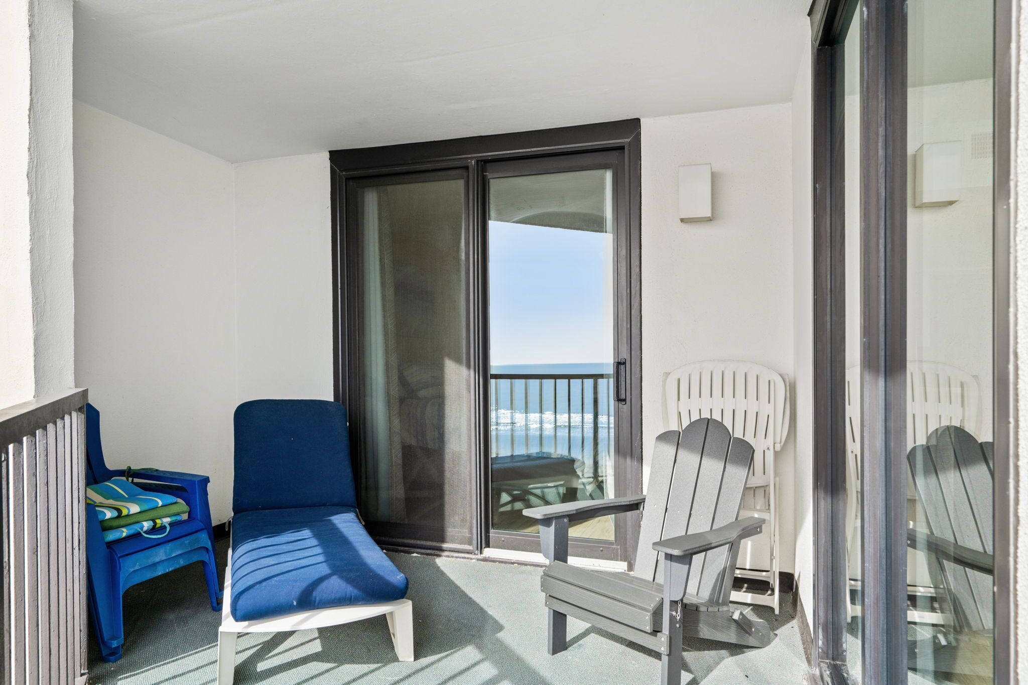 A balcony with a blue lounge chair, a small table with a green and blue cushion, and two Adirondack chairs.