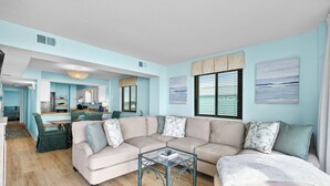 A spacious living room with a beige sectional sofa, a glass coffee table, and a view of the kitchen through an open door. - 10% off weekly stays (Sat-Sat) thru Aug. 3BR 3BA OF condo. Crescent Beach. (North Myrtle Beach)