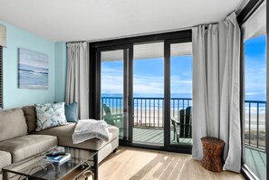 A modern living room with a sofa, a glass coffee table, and a view of the beach through large sliding glass doors. - 15% off 3-7 nights thru Nov. Oceanfront condo. Crescent Beach. Amazing views! (North Myrtle Beach)