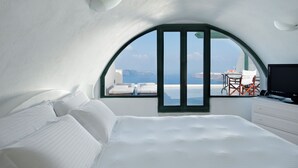 A modern bedroom with a large bed, a view of the sea, and a small table with a chair. - Anastasis Apartments and Spa (Santorini)