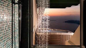 A bathroom with a showerhead, a window, and a view of the sea at sunset. - Anastasis Apartments and Spa (Santorini)