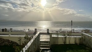 Een uitzicht op zee, met de zon door de wolken schijnend, een trap die naar een gebouw leidt en een hekwerk met uitzicht op het water.