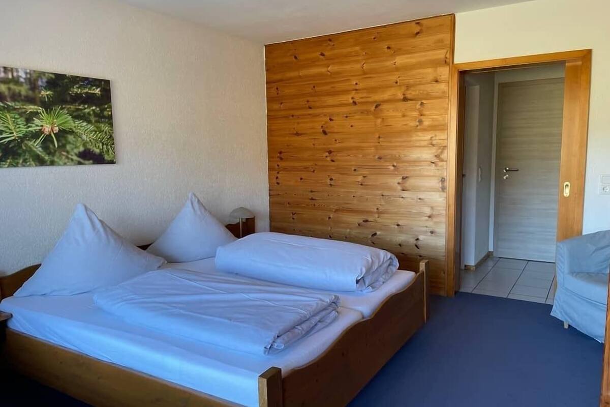 A room with a wooden bed, a wooden panel wall, a door, and a small table with a lamp.