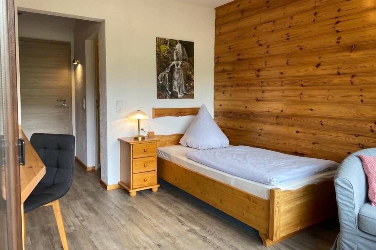 A bedroom with a wooden headboard, a single bed, a nightstand with a lamp, a chair, and a door leading to another room.