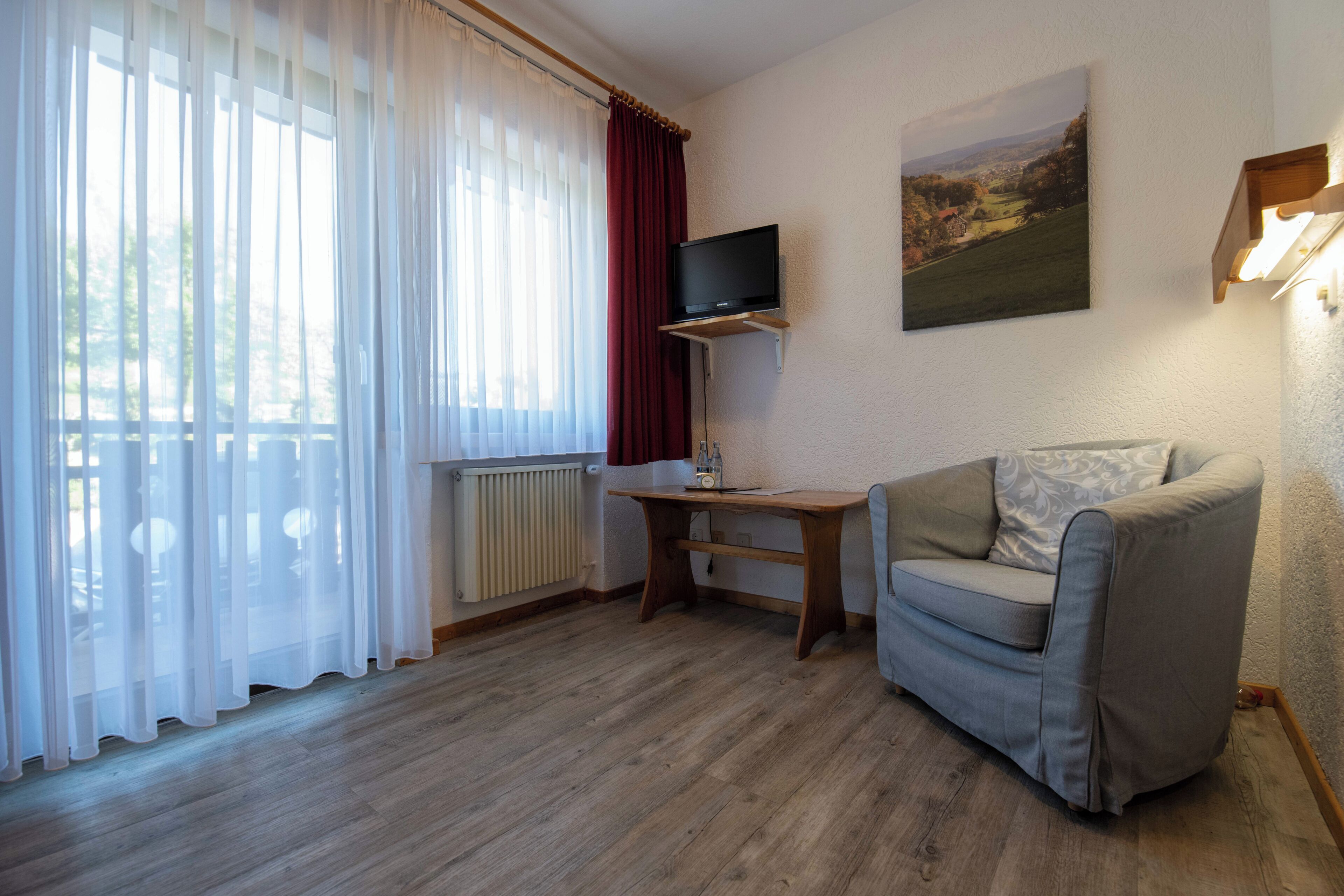 A room with a wooden floor, a grey sofa, a wooden table with a television on top, and a window with sheer curtains.