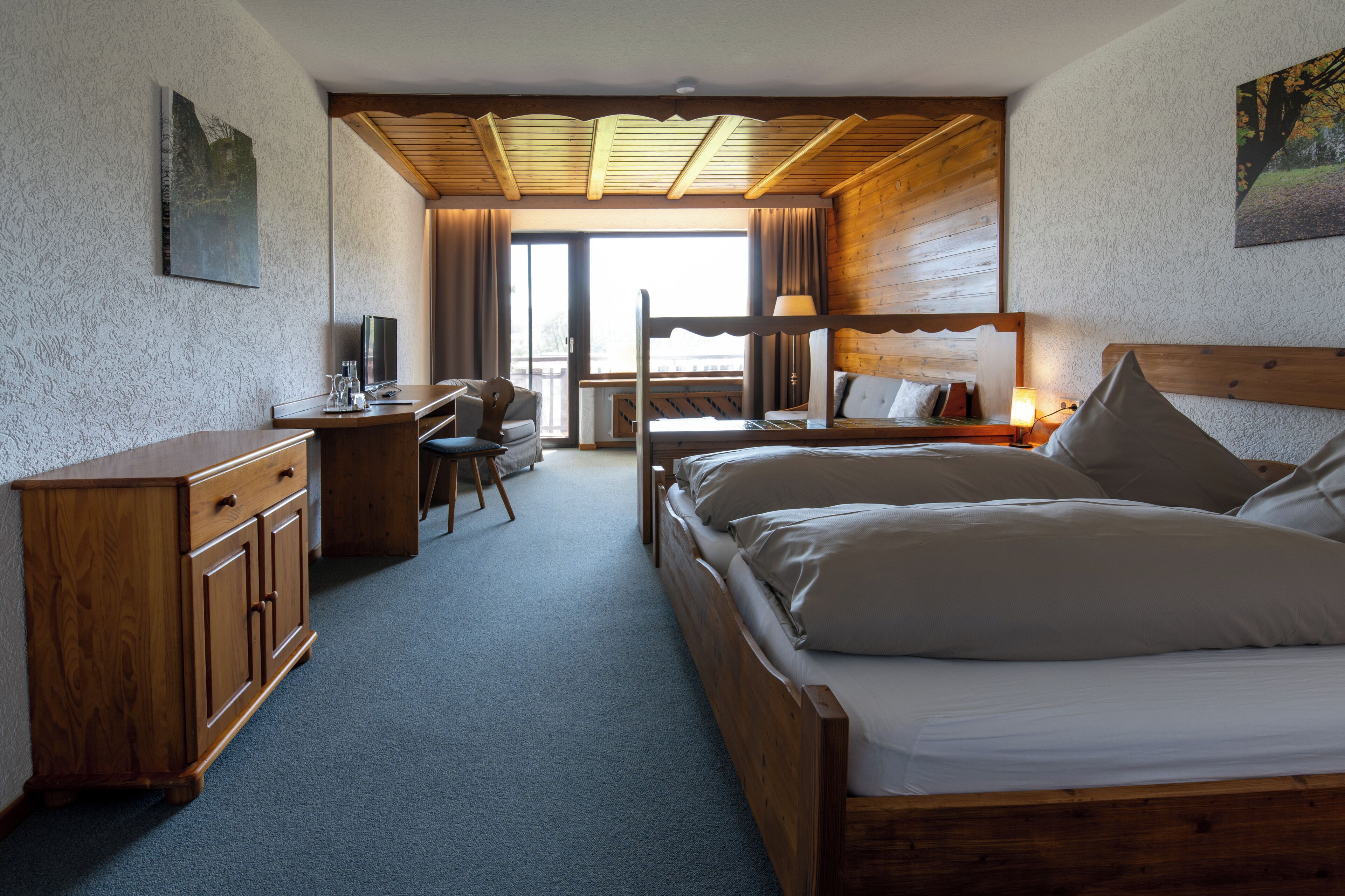 Una habitación de hotel con una cama de madera, un escritorio con una silla, un televisor y un ventanal amplio.