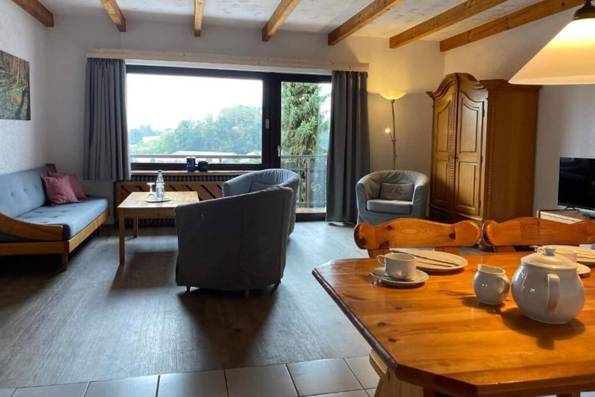 A cozy living room with a sofa, chairs, a wooden table with tea set, and a large window with a view of greenery.