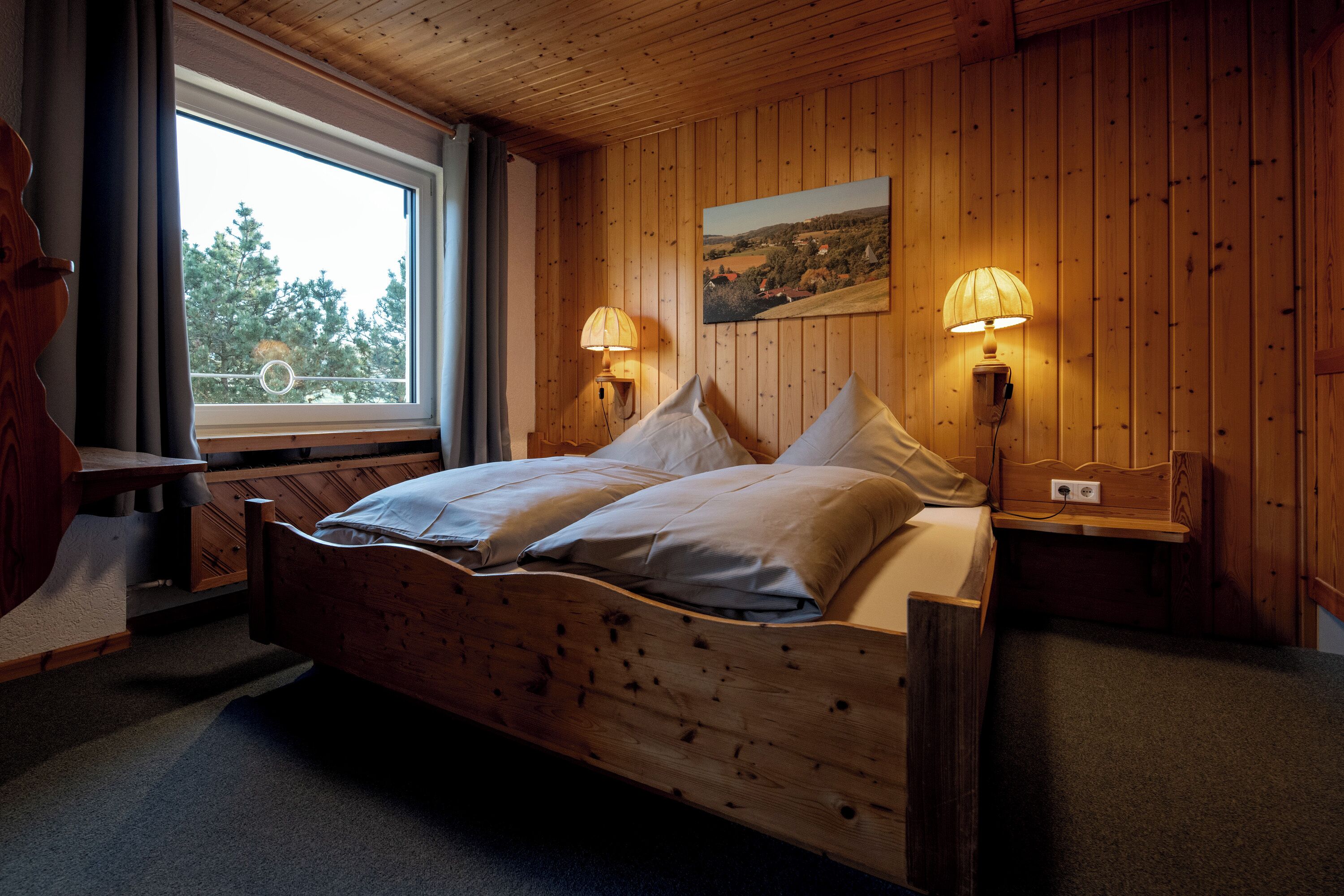 Una habitación con paneles de madera, una cama, dos lámparas de noche, una ventana y un cuadro en la pared.