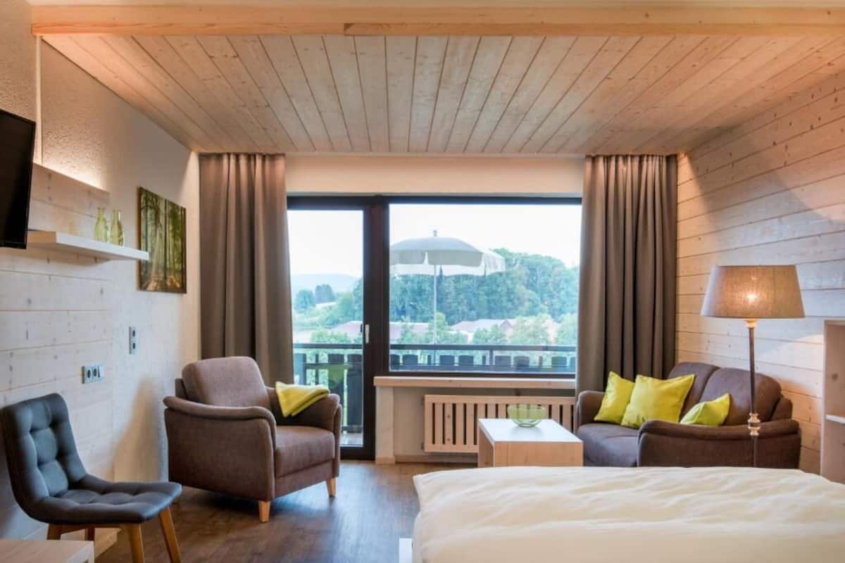 A hotel room with a bed, two armchairs, a small table, a lamp, and a view of a balcony with a railing and greenery.