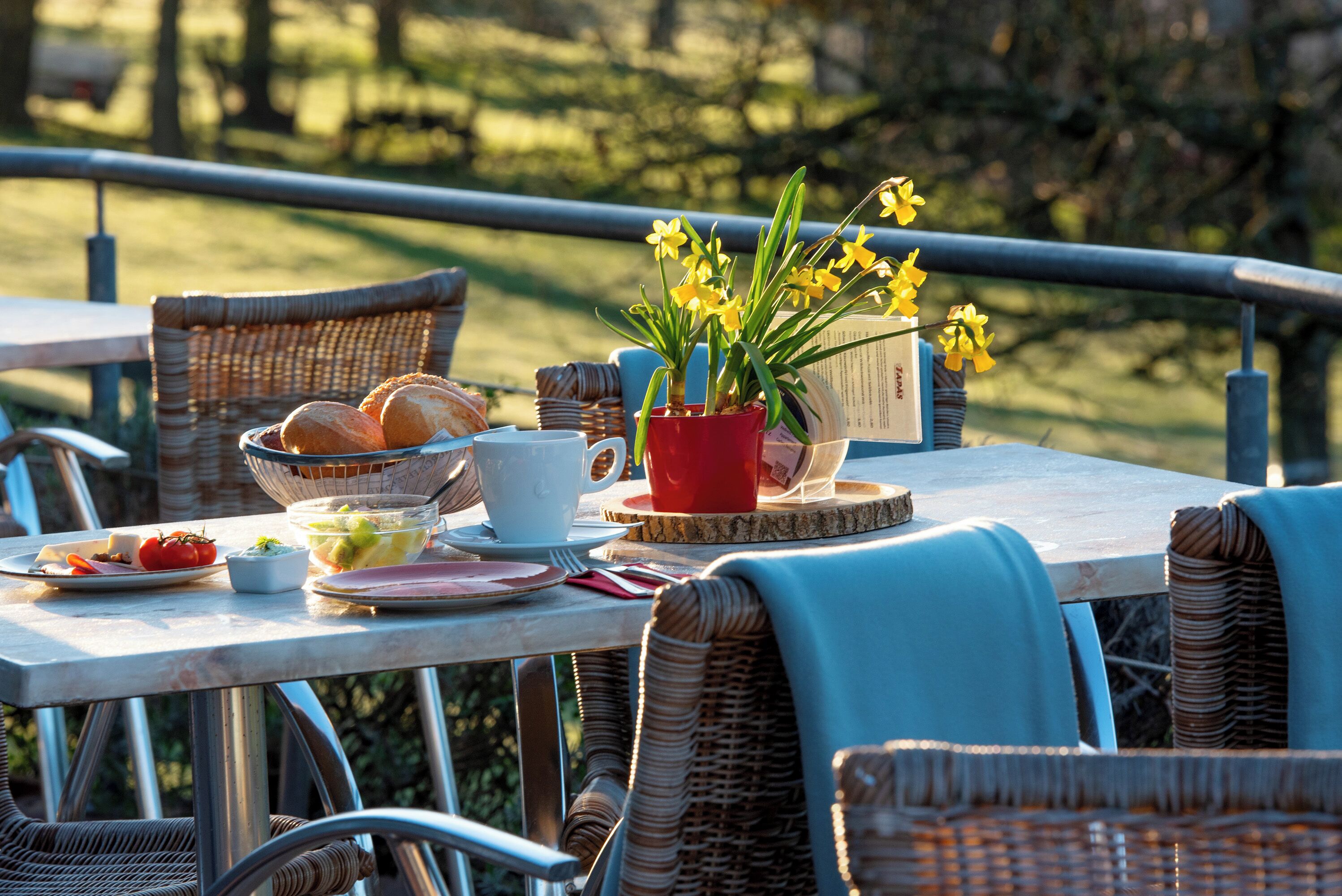 Een terras met rieten stoelen, een tafel gedekt met brood, fruit en een rode bloempot.