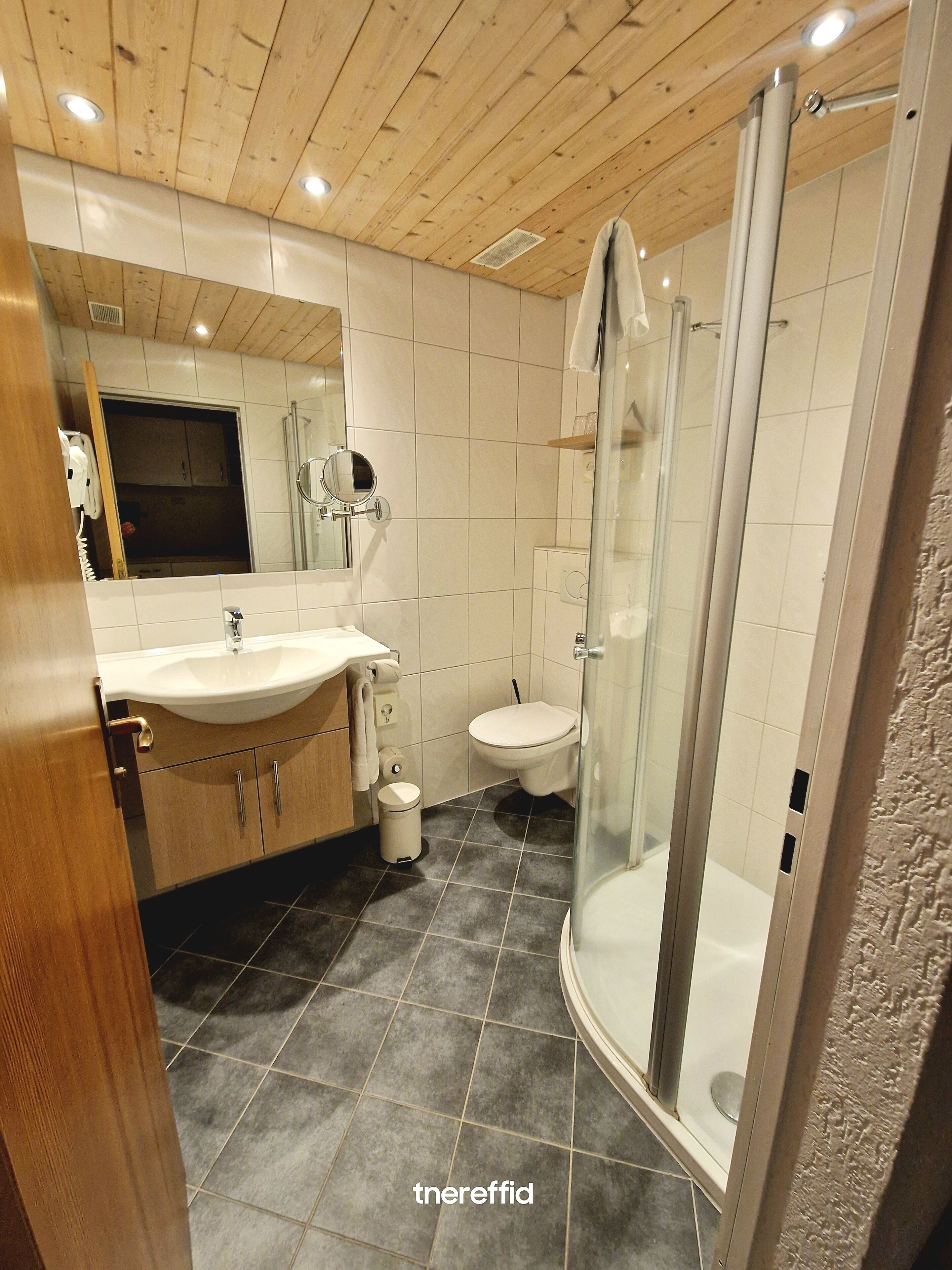 Ett modernt badrum med ett träpanelat tak, ett handfat, en toalett och en dusch med glasväggar.