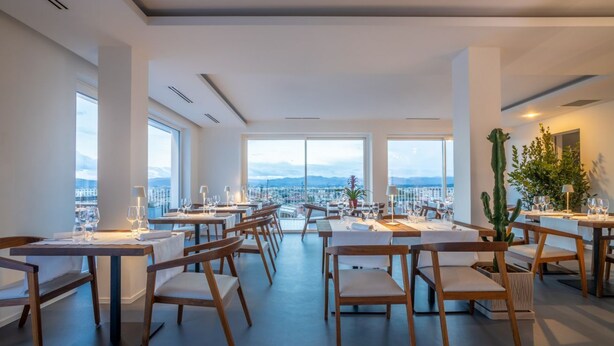En restaurant med borde og stole i et rummeligt, velbelyst interiør. Store vinduer giver udsigt over bybilledet og bjergene.
