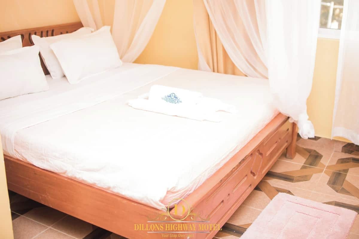 A neatly made bed with white linens and a towel, a wooden headboard, and a view of the outdoors through sheer curtains.