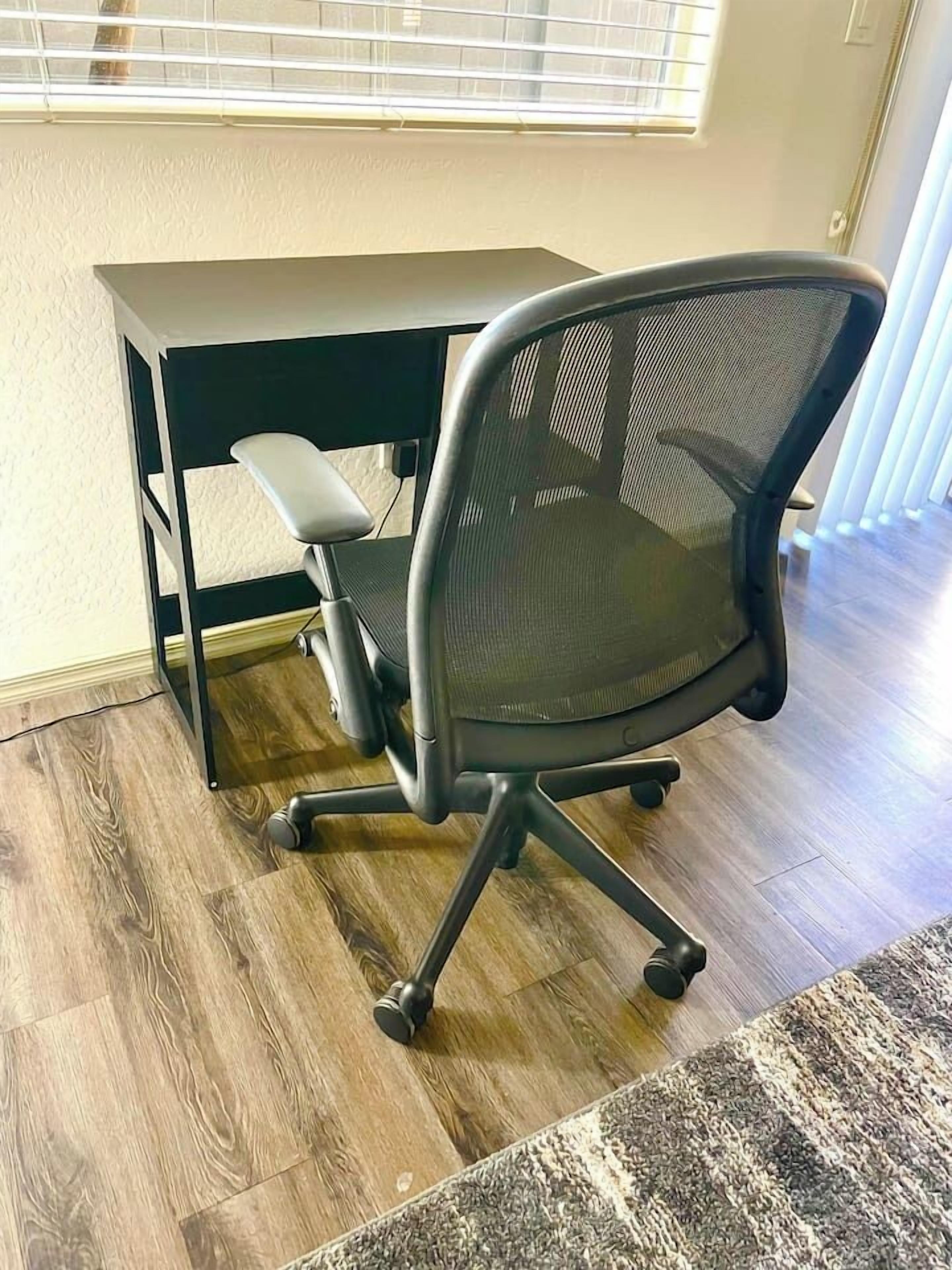 Un fauteuil de bureau moderne doté d’un dossier en tissu à mailles et de roulettes, placé sur un plancher en bois, avec un bureau de couleur sombre en dessous.