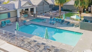 Ein Swimmingpool mit Liegestühlen und Sonnenschirmen, umgeben von Gebäuden im modernen Stil und Palmen.