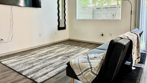 Ein modernes Wohnzimmer mit einem großen Flachbildfernseher an der Wand, einem ungewöhnlichen Wellenspiegel, einem großen Fenster mit Jalousien, einem gemusterten Teppich und einem Sofa mit Decke.